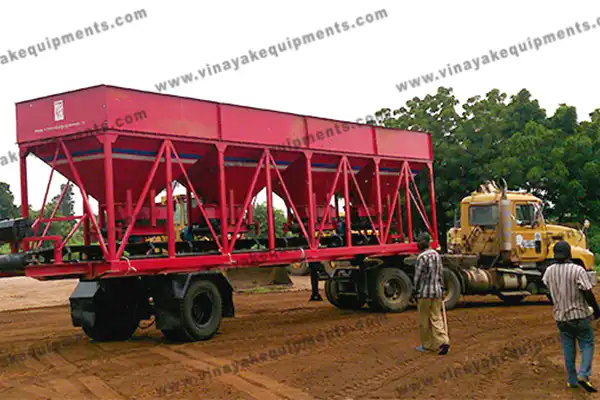 Red steel lattice-frame equipment on a trailer being hauled by a yellow truck, with two men watching on a dirt road area.